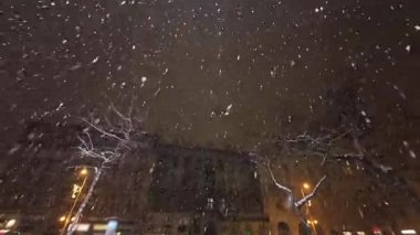 Avrupa şehir binalarının üzerine yağan yoğun kar yağışı ve geceleri sokak ışıklarıyla aydınlatılan çıplak ağaçların geniş açılı görüntüsü.