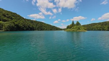 Turkuaz bir tatlı su gölü, küçük bir orman adası ve açık bir gökyüzünün altındaki yeşil tepeleri çevreleyen bir hava videosu. Doğal manzara görüntüleri editoryal stok kullanımı için uygundur.