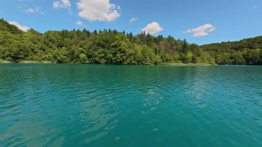 Sakin turkuaz tatlı su gölü videosu. Küçük yüzey dalgaları ve açık mavi gökyüzünün altında karışık bir orman şeridi. Doğal arka plan görüntüsü olarak ticari stok kullanımı için uygundur.