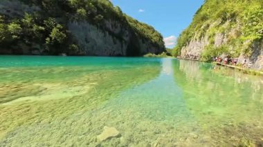 Temiz tatlı su gölü, görülebilir sığ tabanı, kayalık uçurumları ve gün ışığında ağaçtan göl kenarında yürüyen ziyaretçilerin videosu. Turizmi doğal ortamda tasvir etmek için editoryal stok kullanımına uygundur.