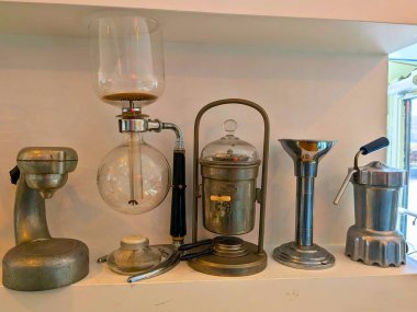 Beyaz bir rafta sergilenen eski kahve demleme ekipmanları koleksiyonu. Retro metal kahve makineleri, cam sifon birası, el değirmeni ve doğal ışık altında kahve makineleri. Kahve kültürü kavramı, geleneksel demleme yöntemleri, işçilik.