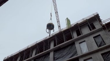 İnşaat halindeki çok katlı bir binanın önündeki vinçte büyük bir endüstriyel beton kova asılı duruyor. Yapının ön cephesi, sarı bir enkaz bacasının yanında siyah koruyucu plastik kaplamayla kaplı..