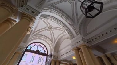 Krem renkli neoklasik sütunların ve dekoratif pervazlı kubbeli tavanın olduğu büyük salonun iç manzarası. Geniş kemerli bir kapı, temiz bir gökyüzünün altında pembe bir cephe binası olan bir avluya açılıyor..