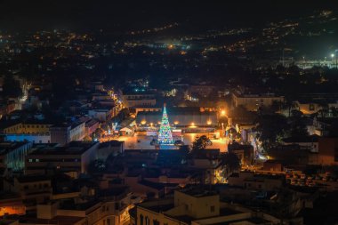 Bir meydanın ortasında aydınlanmış bir çam ağacının ve etrafındaki evlerin olduğu Noel sahnesi