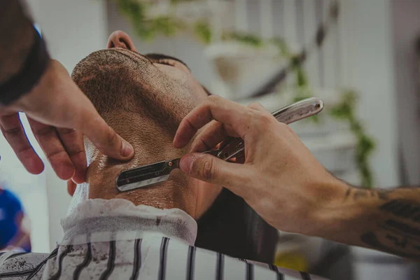 detail of a young man with tattooed arms cuts a man\'s hair in a barber shop