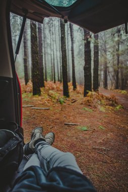 Tarlanın ortasında bir kamp aracının içinde dinlenen bir kişinin ayakları görülüyor, arka planda büyük ağaç gövdeleri görülüyor.