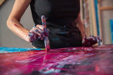 Genç bir kadın, elleri stüdyoda, fırçaya odaklanmış bir şekilde soyut bir resim çiziyor.