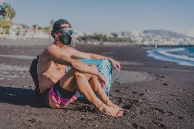 Şapkalı, gözlüklü, sörf kıyafetli genç adam maske takmış denize bakıyor.