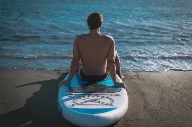 Genç bir sporcu, mavi gökyüzünün altında sahilde sörf yaptıktan sonra dinleniyor.