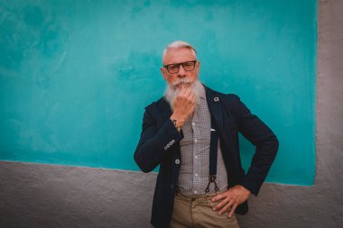 Hipster elbiseli, gözlüklü ve uzun beyaz sakallı yaşlı bir adam mavi bir duvarda duruyor.