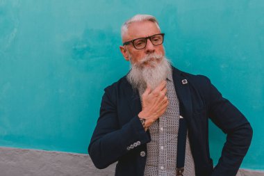 Hipster elbiseli, gözlüklü ve uzun beyaz sakallı yaşlı bir adam mavi bir duvarda duruyor.