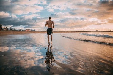 Genç bir adam plajda gün batımında egzersiz yapıyor. Sıcak renkler