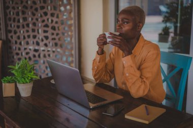 Siyah yetişkin bir kadın kahve içerken bir kafede bilgisayarının başında yüzüne odaklanıyor.
