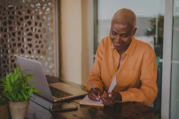 黑人成年妇女一边在咖啡店的电脑上工作 一边在书上写着便条 图库照片