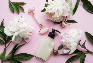 Yüz masajı terapisi, masaj yağları ve pembe şakayıklar için yeşim bir yüz silindiri. Pembe pastel arka planda düz uzandı