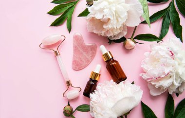 Yüz masajı terapisi, masaj yağları ve pembe şakayıklar için yeşim bir yüz silindiri. Pembe pastel arka planda düz uzandı