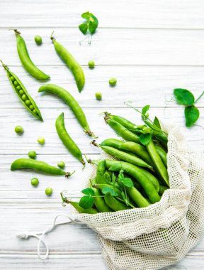 Beyaz ahşap arka planda taze bezelye kabuklarıyla dolu yeniden kullanılabilir çevre dostu çanta.