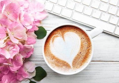 Pembe ortanca çiçeği buketli ofis masası çalışma alanı, beyaz ahşap arka planda bir fincan kahve ve klavye. Flatlay, üst görünüm.