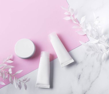 Mermer ve pembe arka planda beyaz krem tüpleri ve beyaz yapraklı dekoratif dallar.