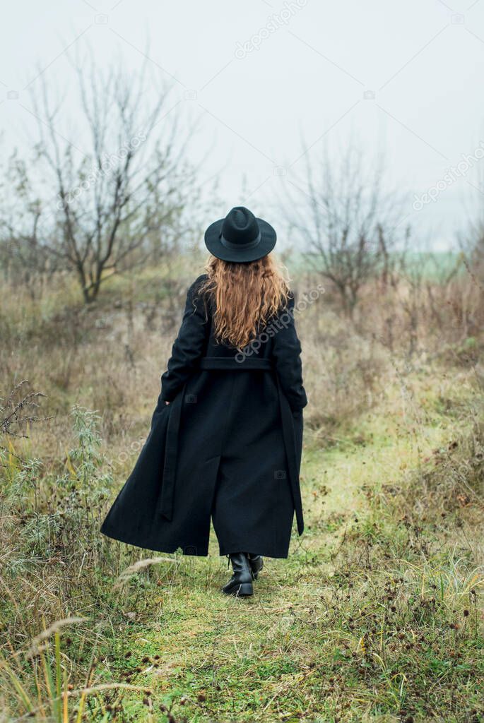 Portrait of young attractive woman in black coat and hat. Autumn landscape, dry grass. Back vie