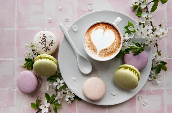 Macaroons with a cup of coffee and a branch of white flowers on a pink tile background. French dessert and flowers