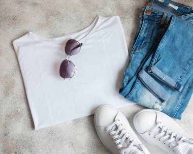 Şık kadın kıyafetleri seti. Beton zeminde kadın kıyafeti. Mavi kot, beyaz tişört, beyaz spor ayakkabı ve güneş gözlüğü. Düz yatış, üst görünüm