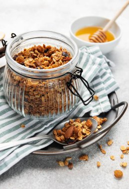 Granola taneleri, fındıklar ve kurutulmuş meyveler gri arka planda ballı bir kavanozda.