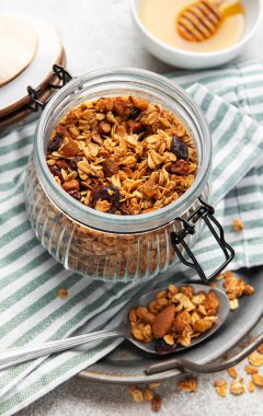 Granola taneleri, fındıklar ve kurutulmuş meyveler gri arka planda ballı bir kavanozda.