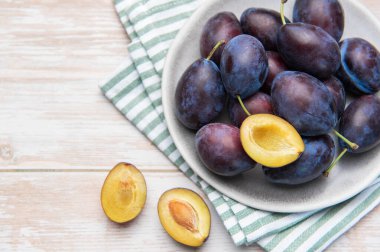 Fresh ripe plums in a bowl, with halved plums on a light wooden background