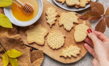 Hand holding a leaf shaped cookie among other autumn inspired shortbread and a bowl of honey