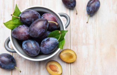 Fresh ripe plums and green leaves filling a grey bowl on a light wooden table