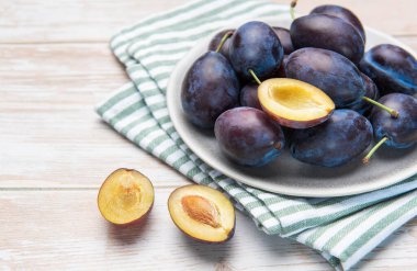 Fresh ripe plums in a bowl, with halved plums on a light wooden background
