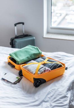 Parlak bir otel odasındaki beyaz bir yatakta düzgün katlanmış giysilerle dolu bavul.