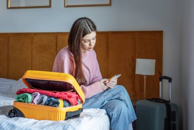 Bir kadın açık bavulla yatakta oturuyor, seyahat ederken akıllı telefon kullanıyor.