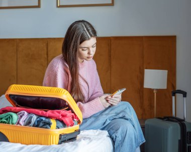 Bir kadın açık bavulla yatakta oturuyor, seyahat ederken akıllı telefon kullanıyor.