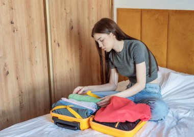 Yatakta oturan bir kadın yatak odasında açık sarı bir bavula giysiler yerleştiriyor.