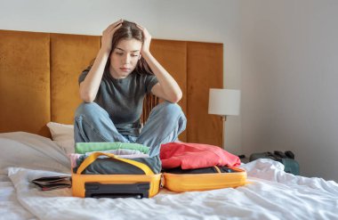 Genç bir kadın yatakta açık bir bavula giysilerini yerleştirirken hayal kırıklığına uğramış hissediyor.