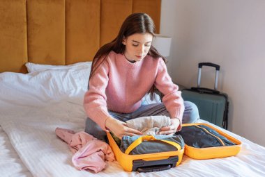 Genç bir kadın beyaz bir otel yatağında açık bir valiz çantasına kıyafet yerleştiriyor.