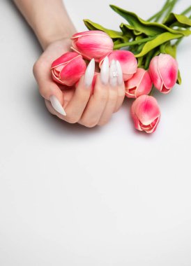 Elinde pembe lale çiçekleri olan kristallerle süslenmiş beyaz topuklu bir kadın eli.