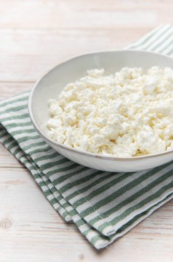 Açık ahşap arka planda yeşil çizgili bir peçeteyle beyaz bir kasede süzme peynir.