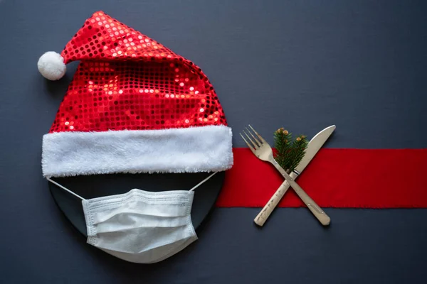 Noel konsepti masası. Tıbbi maskeli bir Noel yemeği. Coronavirus ve Noel 2020 masa konsepti. Yüz koruyucu maske, Noel Baba şapkası ve xmas şenlik masası dekorasyonu, çatal, bıçak ve ağaç dalları.