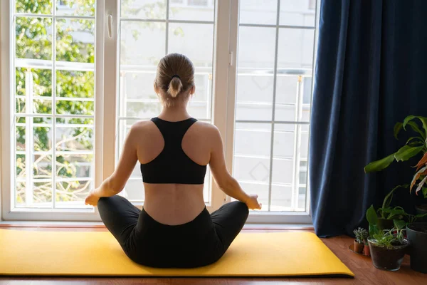Genç bir kadın pencerenin önünde yoga meditasyonu yapıyor. Kısa saç. Tencere ve bitkiler. Sarı yoga minderi. Perde. Rahatlatıcı, ev hobileri, ev karantinası, tecrit, koronavirüs. spor sutyeni