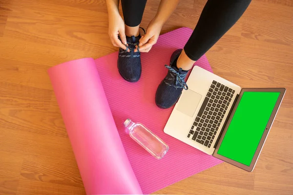 Bağcıklarını bağlayan kadın, spor için hazırlan. Pembe mat su şişesi. Yeşil ekranlı dizüstü bilgisayar. Çevrimiçi yoga meditasyon egzersizi. Koronavirüsü karantinaya alın. Aktif ol. sağlıklı yaşam
