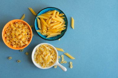 Penne, farfalle tonde kelebeği ve açık mavi arka planda ditalini tipi makarna. Seramik kasede makarna. Mavi turuncu beyaz kase. Soyut, makarna. Üst görüntü fotoğrafı. Dağılmış makarna Kapat. boş