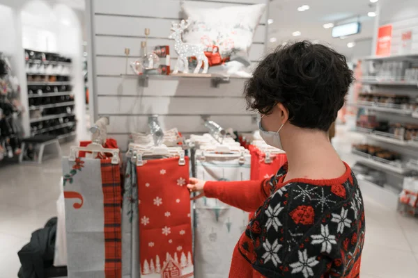 Tıbbi maske takan bir alışveriş kadını. Noel ve yeni yıl alışverişi. hediye fikirleri. Masa örtüsü ve dekorasyon. çirkin Noel süveteri. 2021