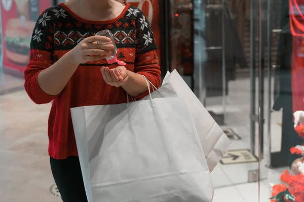 Alışveriş yaparken küçük taşınabilir antibakteriyel dezenfektanı kullanan kadın görüntüsünü kapat. Alışveriş torbasını tut. Noel ya da yeni yıl için. çirkin Noel süveteri. Koronavirüs salgını. 2021