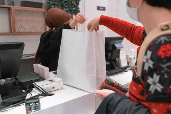 Maske takan kadın alışverişini tamamlıyor. Parasını öder ve çantasını geri alır. Başörtüsü, türban, türban ve maske takan kasiyer kadın alışveriş çantasını uzatıyor. çeşitlilik. Müslüman kadın..