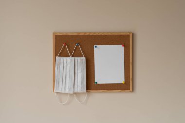 Cerrahi ve tıbbi maskeler beyaz arka planda ahşap çerçeveli bir mantar tahtada renkli raptiyelerde asılı. Coronavirus salgını 2021, kişisel bakım, hatırlatıcı boş kağıt, notlar, uyarı, kopyalama alanı