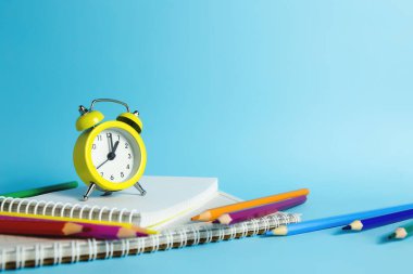 Mekanik alarm saati, defter, mavi arka planda kalemler.