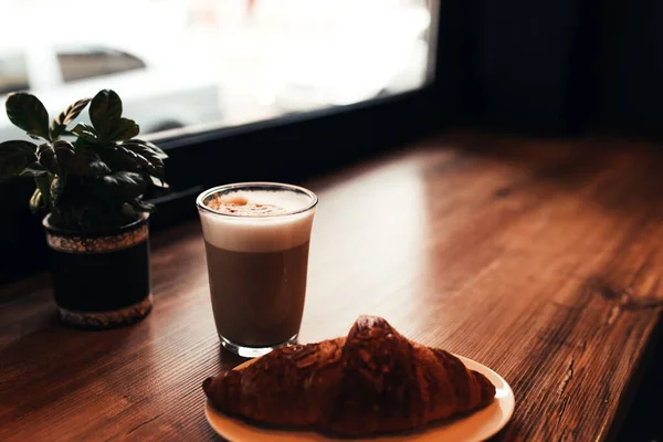 Secangkir kopi, croissant di meja dekat jendela di kafe. Latar belakang kabur — Stok Foto